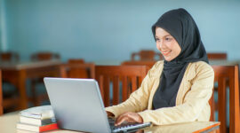 yogyakarta, indonesia february 09, 2023
beautiful asian young woman wearing hijab smiling at work, typing operating laptop computer and mobile phone at her desk.