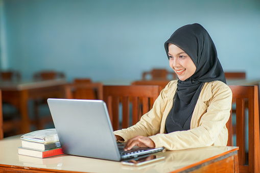 yogyakarta, indonesia february 09, 2023
beautiful asian young woman wearing hijab smiling at work, typing operating laptop computer and mobile phone at her desk.