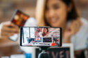 Happy young attractive Asian ethnic model filming vlog on mobile phone on tripod and showing eyeshadows at table with cosmetics