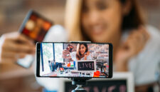 Happy young attractive Asian ethnic model filming vlog on mobile phone on tripod and showing eyeshadows at table with cosmetics