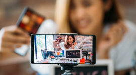 Happy young attractive Asian ethnic model filming vlog on mobile phone on tripod and showing eyeshadows at table with cosmetics