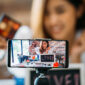 Happy young attractive Asian ethnic model filming vlog on mobile phone on tripod and showing eyeshadows at table with cosmetics